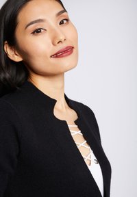 Cardigan noir en tricot avec un col festonné et un détail à laçage blanc à l'avant. Texture lisse avec un design ajusté.