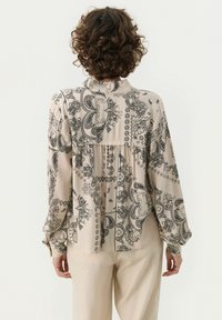Ljust beige blus med svart paisleymönster, långa puffärmar och rundad krage. Har en plisserad design bak för en avslappnad passform.