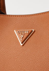 Brown textured leather handbag featuring a triangular gold and brown Guess USA logo emblem on the front.