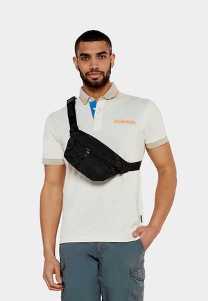 Borsa a tracolla nera indossata a crossbody su una polo bianca con accenti beige e blu. La borsa presenta un materiale texturizzato e una chiusura lampo.