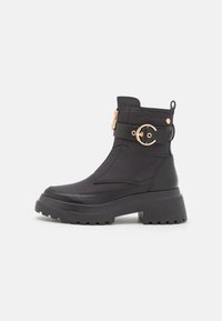 Botas negras hasta el tobillo con suela gruesa, parte superior de textil, acentos en dorado y cremallera lateral. Presenta un detalle de correa con hebilla.