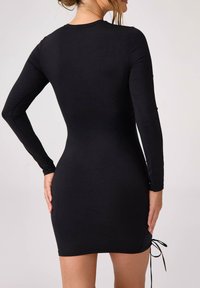 Czarna sukienka z długim rękawem o dopasowanym kroju, wykonana z gładkiego materiału, z detalem sznurków u dołu, umożliwiającym regulację długości.