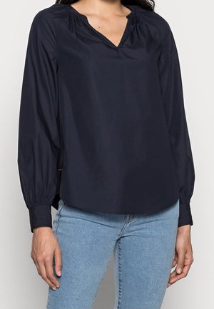 Kobieta ubrana w ciemnogranatową bluzkę z długim rękawem z marszczonymi mankietami oraz jasnoniebieskie dżinsy, stojąca na jednolitym tle.