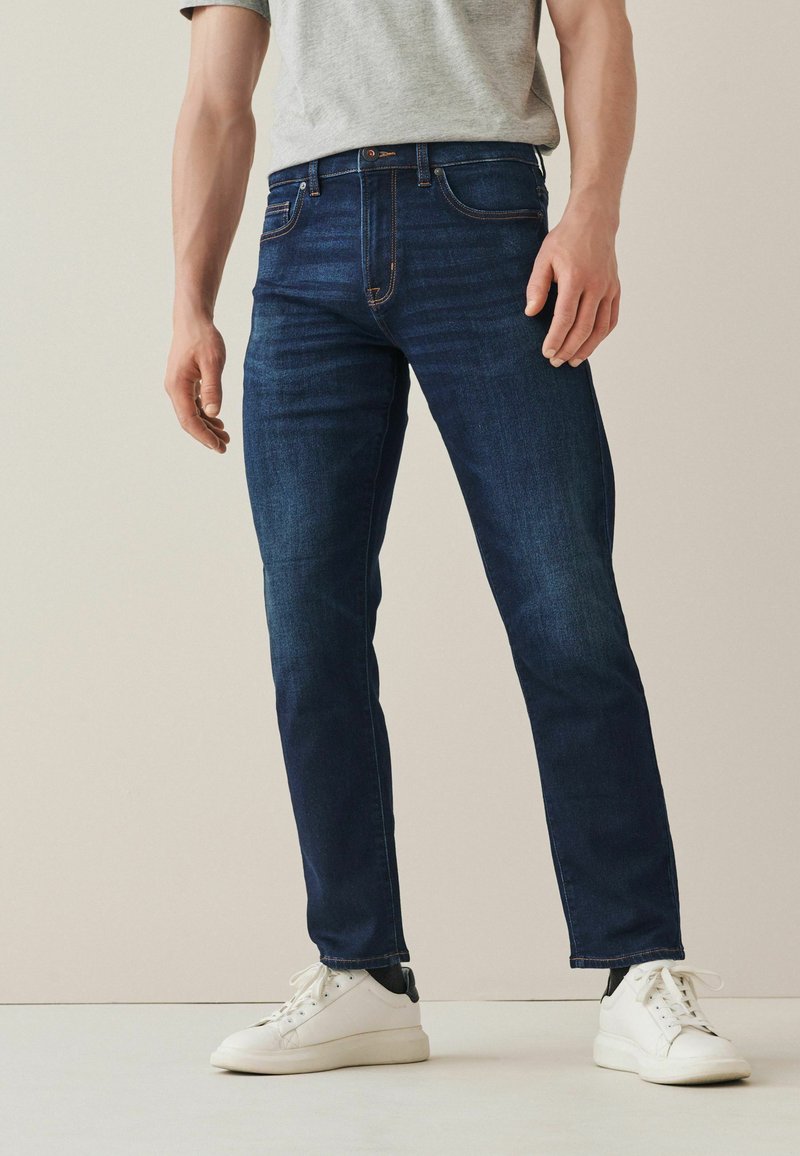 Donkerblauwe jeans met een rechte pasvorm, voorzien van vijf zakken, oranje stikselaccenten en gecombineerd met witte sneakers.