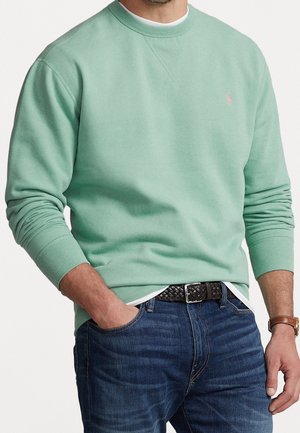 Homme portant un sweat-shirt vert clair à col rond, un jean bleu avec une ceinture tressée marron, un sous-vêtement blanc et une montre en cuir marron.