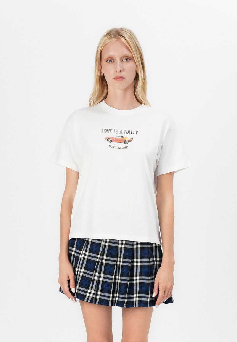White cotton t-shirt with short sleeves and round neckline, featuring printed text and a graphic of a car. Paired with a blue plaid skirt.