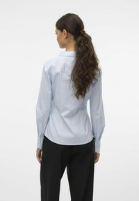 Camisa de manga larga azul claro, hecha de tela suave, con un cuello puntiagudo, dobladillo curvado y puños con botones, combinada con pantalones negros.