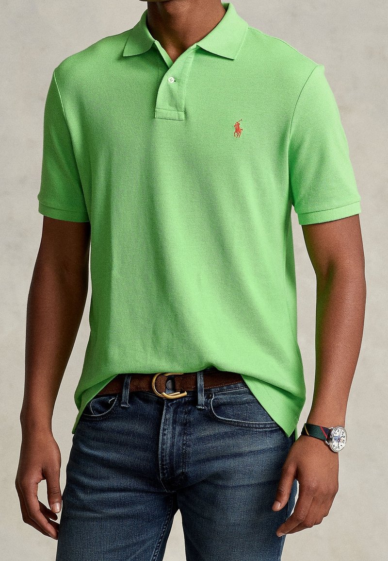 Homme portant un polo vert vif avec un petit logo rouge, un jean bleu foncé, une ceinture marron et une montre-bracelet avec un bracelet foncé.