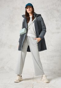 Manteau isolé gris en tissu texturé, superposé à un pull gris clair et un pantalon de jogging large. Une casquette teal et un petit sac menthe complètent le look.