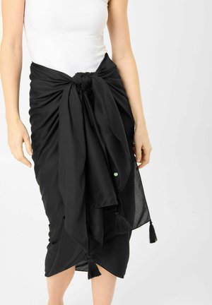 Vrouw draagt een zwarte sarong, gebonden in de taille over een wit mouwloos topje, met kwastjes die aan de randen van de sarong hangen.