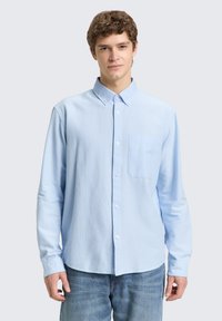 Ανοιχτό γαλάζιο πουκάμισο με μακριά μανίκια και κουμπιά, με γιακά, τσέπη στο στήθος και κουμπότρυπες. Φορεμένο με τζιν παντελόνι.