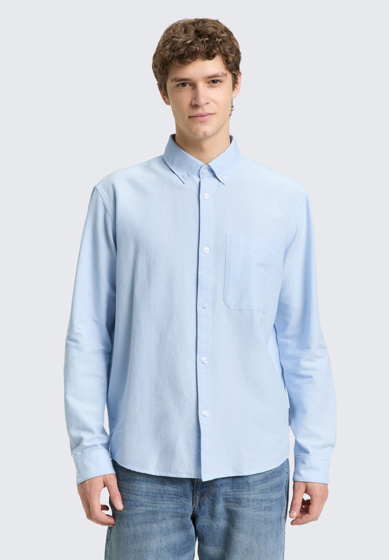 Ανοιχτό γαλάζιο πουκάμισο με μακριά μανίκια και κουμπιά, με γιακά, τσέπη στο στήθος και κουμπότρυπες. Φορεμένο με τζιν παντελόνι.