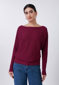 Μπορντό πλεκτό πουλόβερ με ραβδώσεις, φαρδύ καραβόσχοινο λαιμόκοψη και μακριά μανίκια. Συνδυασμένο με μπλε ψηλόμεσα τζιν παντελόνια με πίσω τσέπες.