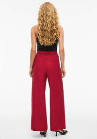 Brede rode broek met hoge taille, zachte stof en riemlussen. Model draagt een zwart mouwloos topje en zwarte hakken.