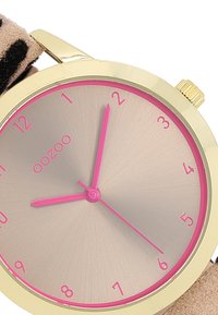 Montre ronde avec un boîtier doré, index des heures et aiguilles roses. Elle possède un bracelet en tissu imprimé animal beige.