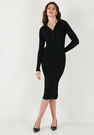 Frau, die ein enges, langärmliges schwarzes Ripp-Midikleid mit Kragen und Knöpfen trägt, kombiniert mit schwarzen spitzen Pumps mit Kettendetails.