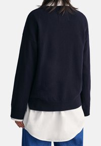 Marineblauer Pullover mit lockerem Sitz, strukturiertem Strickmuster und geripptem Saum. Kombiniert mit einem weißen, geschichteten Hemd, das einen lässigen, zeitgemäßen Stil zeigt.