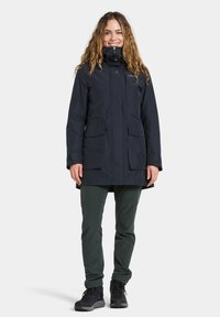 Woman with long curly hair wears a navy waterproof coat, dark green pants, and black hiking boots, standing against a plain background.