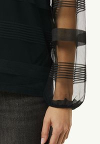 Schwarzoberteil mit durchsichtigen gestreiften Netzärmeln und elastischen Bündchen. Hervorstechende Texturvariationen zwischen soliden und transparenten Paneelen.