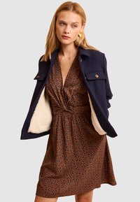 Giacca in misto lana blu navy con fodera crema e dettagli con bottoni indossata sopra un vestito marrone con piccoli motivi floreali blu e vita cinchata.