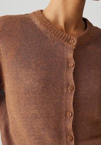 Zbliżenie na osobę noszącą brązowy sweter zapinany na guziki z prążkowanym kołnierzem na jednolitym tle.