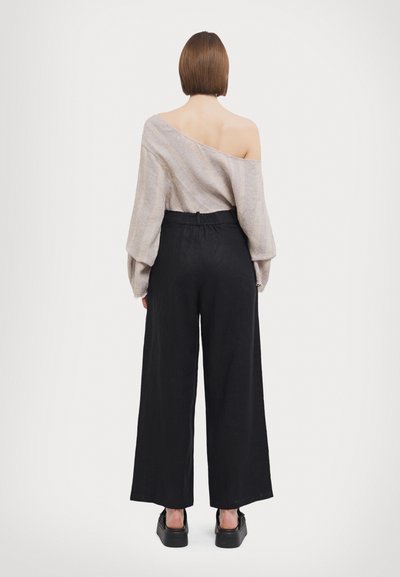 Opuščen bež top s spuščenimi rameni in teksturirano tkanino, v kombinaciji z visokim pasom črnimi širokimi hlačami in črnimi platform čevlji, pogled od zadaj.