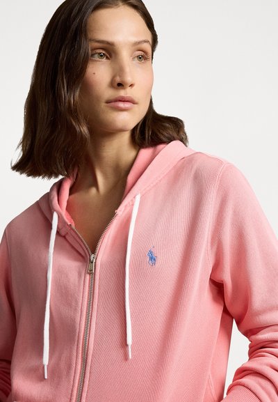 Casaco com fecho éclair rosa em tecido macio e texturizado, com cordões brancos e pequeno logo azul no peito esquerdo. Design casual, forma ajustada.
