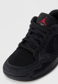 Zapatilla Air Jordan negra con logo Jumpman rojo en la lengüeta, paneles de malla y ante, y cordones negros sobre un fondo blanco.