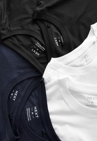 Négy póló fekete, tengerészkék és fehér színekben. Puha pamutból készült, kerek nyakkivágással és "NEXT" feliratos, "Regular Fit" jelzésű címkékkel.