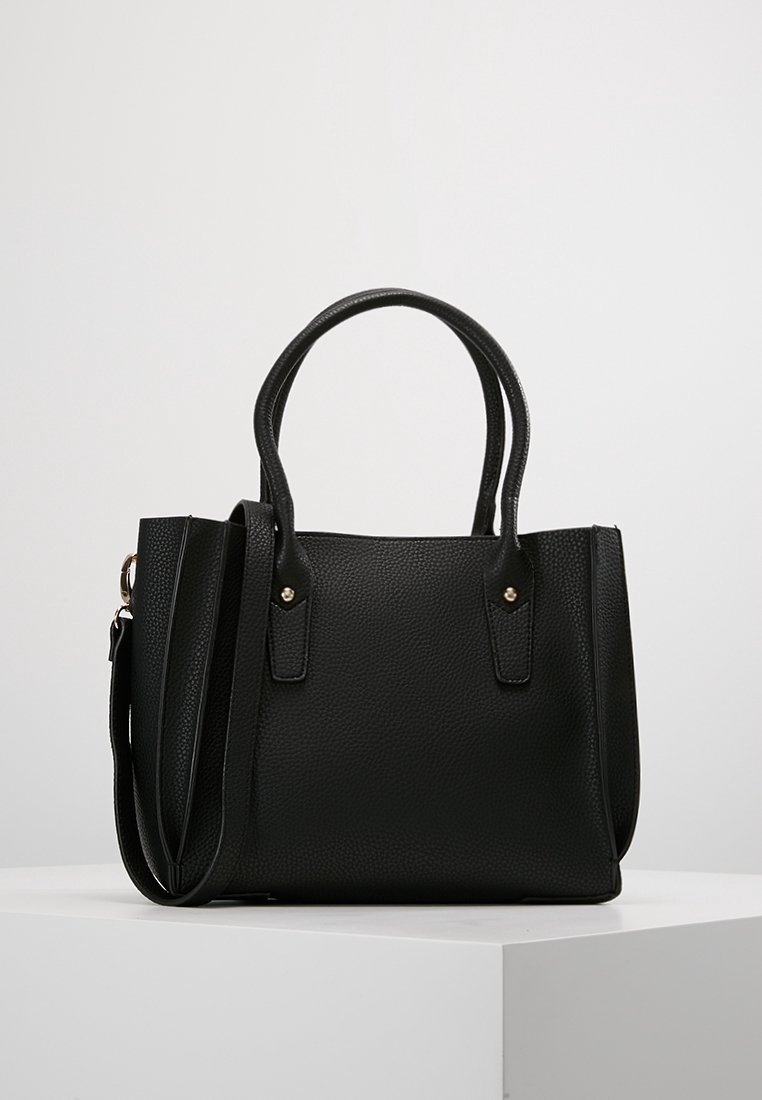Sac à main en cuir noir texturé avec deux poignées supérieures et une bandoulière amovible sur un socle blanc.