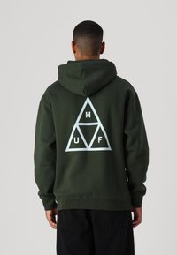 Sudadera verde oscura con un gran logo triangular blanco en la parte posterior, que presenta las letras "H", "U" y "F" dentro de los triángulos. Textura de tela suave.