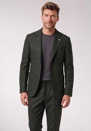 Man with curly hair wearing dark green suit jacket and matching trousers over a dark grey shirt, standing against a plain light background.