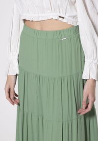 Falda en verde con volantes, hecha de una tela ligera, que presenta una cinturilla fruncida y un pequeño logo metálico cerca de la parte superior.