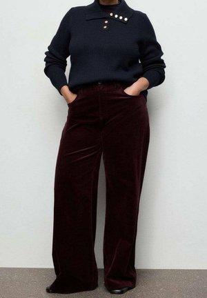 Person trägt einen dunkelmarineblauen, gerippten Pullover mit goldenen Knöpfen und hoch taillierte, weite bordeauxfarbene Cordhosen, Hände in den Taschen.