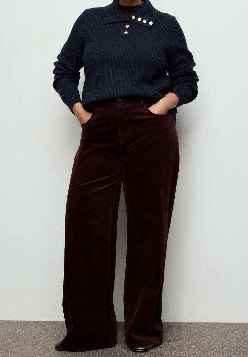 Personne portant un pull côtelé bleu marine foncé avec des boutons dorés et un pantalon en velours côtelé bordeaux taille haute à jambes larges, mains dans les poches.