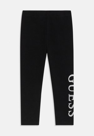 Leggings noirs ajustés avec le texte "GUESS" en blanc pailleté, placé verticalement le long de la partie inférieure externe de la jambe droite.