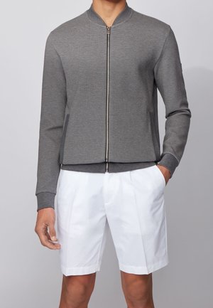 Homme portant une veste bomber grise zippée et un short blanc plissé, debout avec une main dans la poche sur un fond clair.