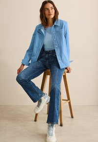 Veste en denim bleu clair avec de grands boutons sur un haut bleu clair, associée à un jean évasé de ton moyen et des baskets blanches avec des accents bleus.