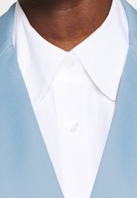 Blazer azzurro chiaro con texture, indossato sopra una camicia bianca con colletto, caratterizzato da un bottone di chiusura al collo e dettagli in tessuto liscio.