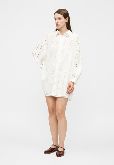 Kvinde iført en løs hvid skjortekjole med lange ærmer og lodrette udskæringsmønstre samt mørkebrune Mary Jane-sko, stående med den ene hånd bag ryggen.