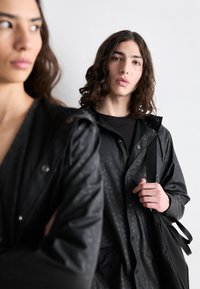 Des vestes de pluie noires avec des motifs texturés, équipées de boutons pression et d'une capuche. Une personne tient un sac noir, tandis que l'autre est de profil.