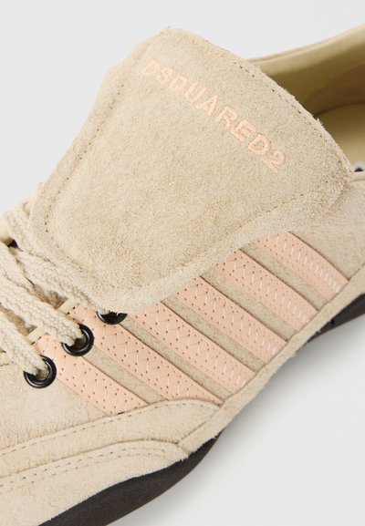 Baskets en suède beige avec un langue rembourrée, des accents rayés roses et des œillets noirs pour les lacets. Semelle en caoutchouc sombre avec surface texturée.