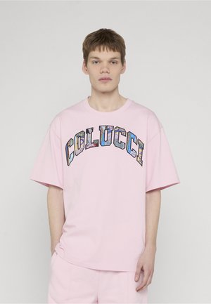 Young man with short brown hair wearing a loose pink T-shirt and shorts set, with colorful "COLUCCI" text on the shirt front.