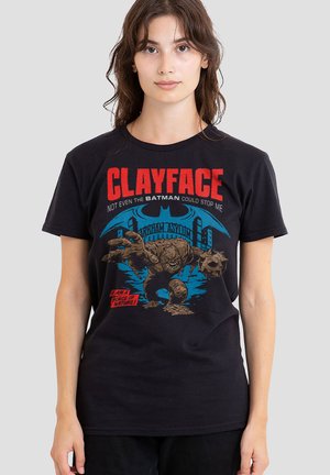 Junge Frau trägt ein schwarzes T-Shirt mit einer Grafik von Clayface aus Batman, die den Bösewicht zeigt, wie er nach vorne greift, mit dem Arkham Asylum im Hintergrund.