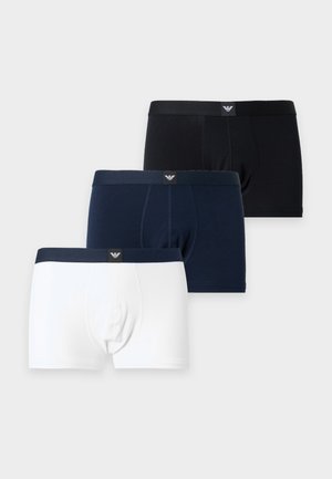 Slanke donkerhuidige mannelijke model staat blootsvoets en draagt zwarte boxershorts tegen een effen lichtgrijze achtergrond.