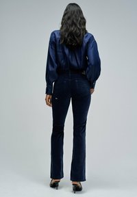 Mujer con cabello oscuro y ondulado, vistiendo una camisa de mezclilla azul oscura, pantalones ajustados azul oscuro a juego y zapatos de tacón negro, de pie mirando hacia atrás.