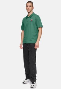 Grön polotröja av bomull med korta ärmar och krage, prydd med en liten logotyp på bröstet, i kombination med svarta jeans och sneakers.