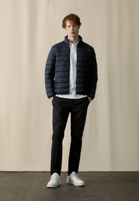 Navy puffer jacket with a high collar and horizontal quilted design, paired with black trousers and white sneakers. Smooth textures visible.