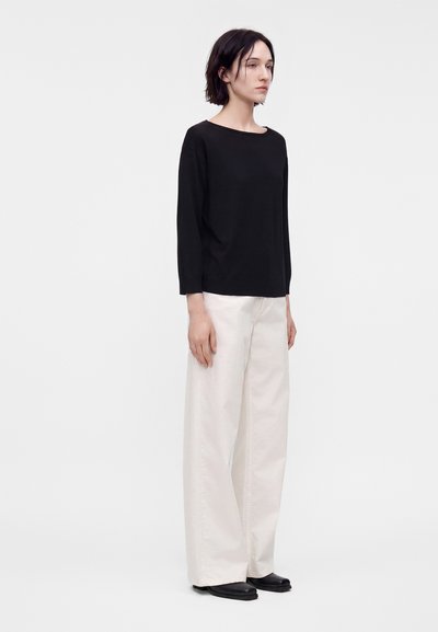 Jeune personne aux cheveux courts et foncés portant un haut noir à manches longues, un pantalon blanc large et des chaussures noires, debout devant un fond clair uni.