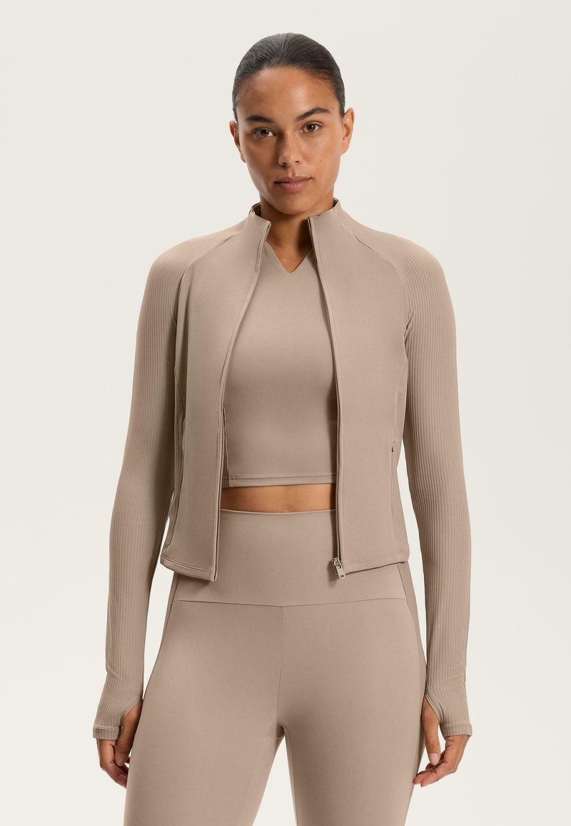 Beige Activewear-Jacke mit vollem Reißverschluss, langen Ärmeln, geripptem Texture und figurbetontem Design, kombiniert mit einem passenden Croptop und Leggings.